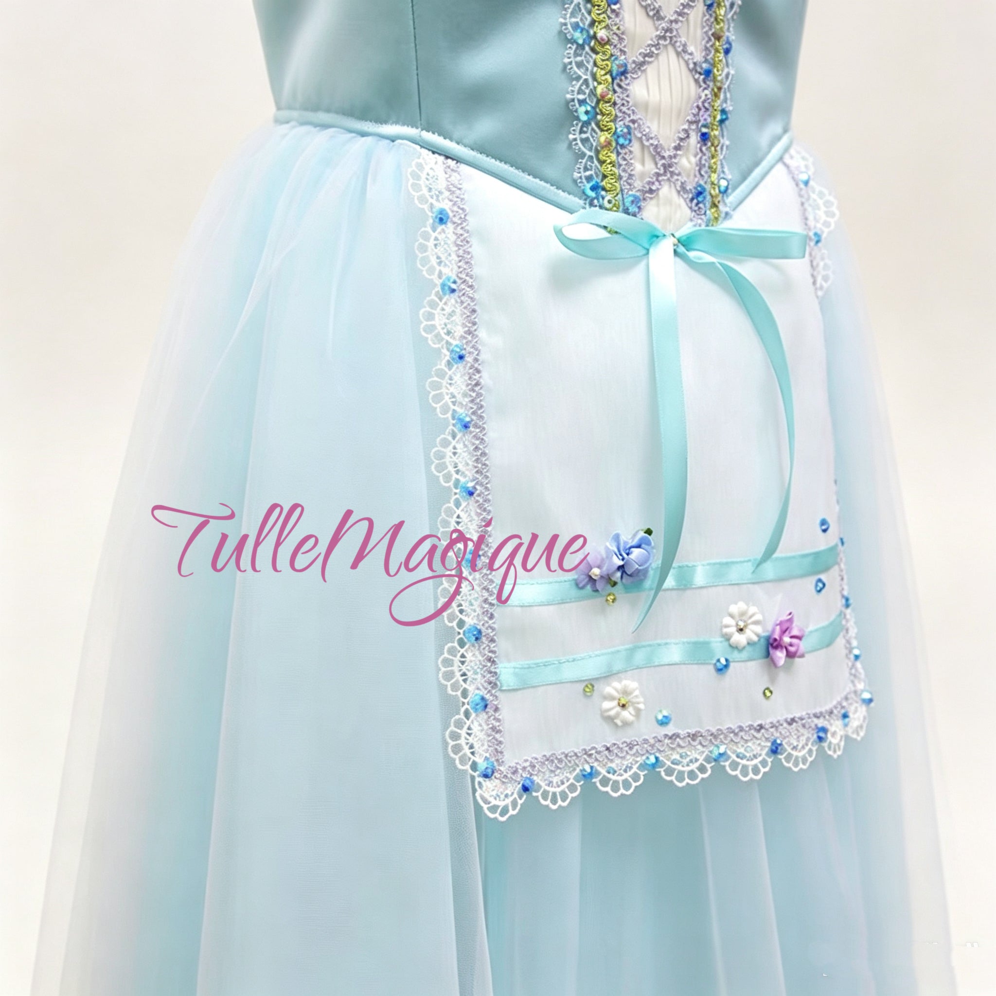 Detailed view of the ballet costume displaying a white lace-trimmed apron with blue ribbons and a flowing tulle skirt with heavy white openwork lace borders.