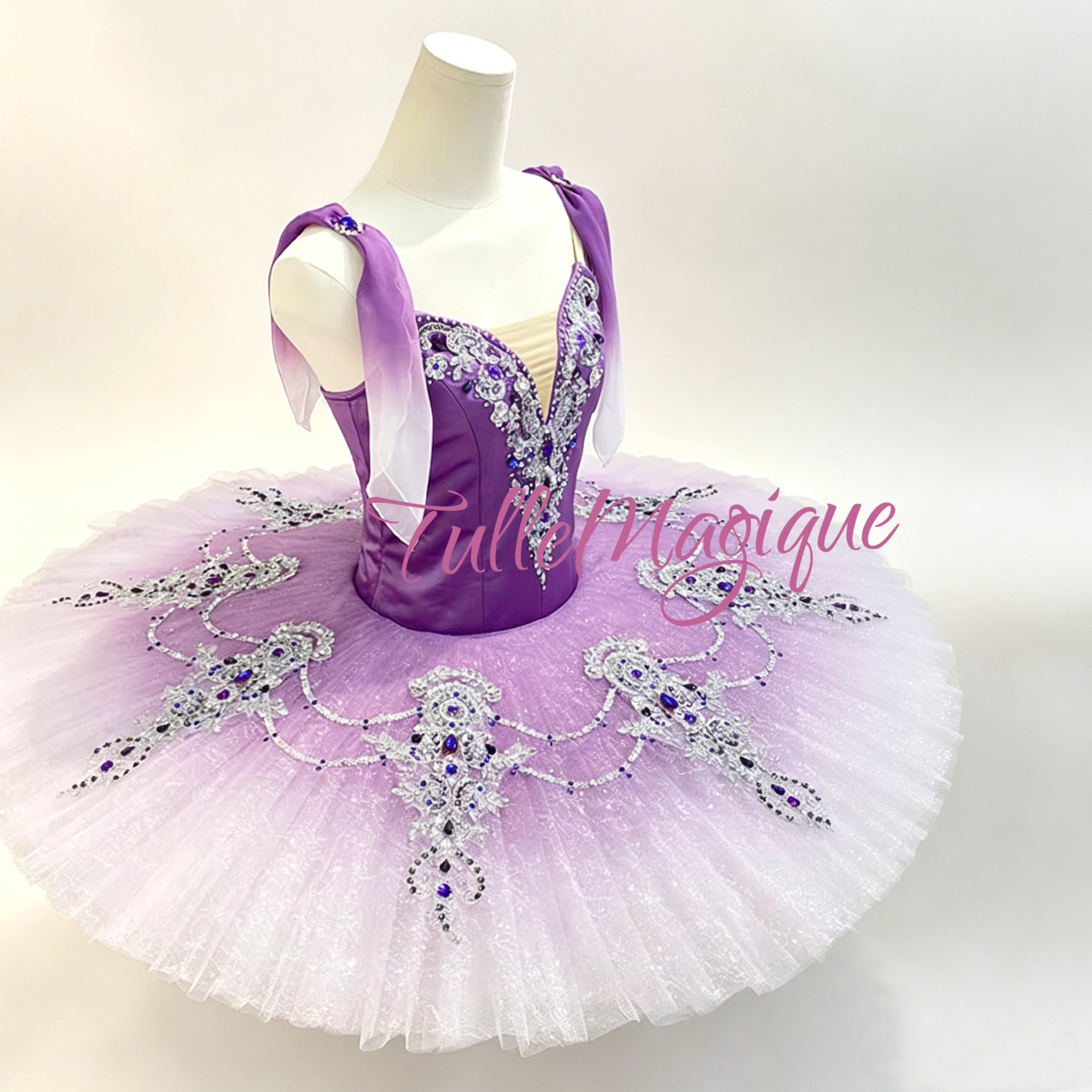 A purple and lavender ballet tutu featureing a purple satin bodice with silver rhinestone embellishments, a lavender sash, and a voluminous white tulle skirt with matching purple and silver rhinestone decorations. This design is perfectly curated for the role of Gulnara in Le Corsaire.